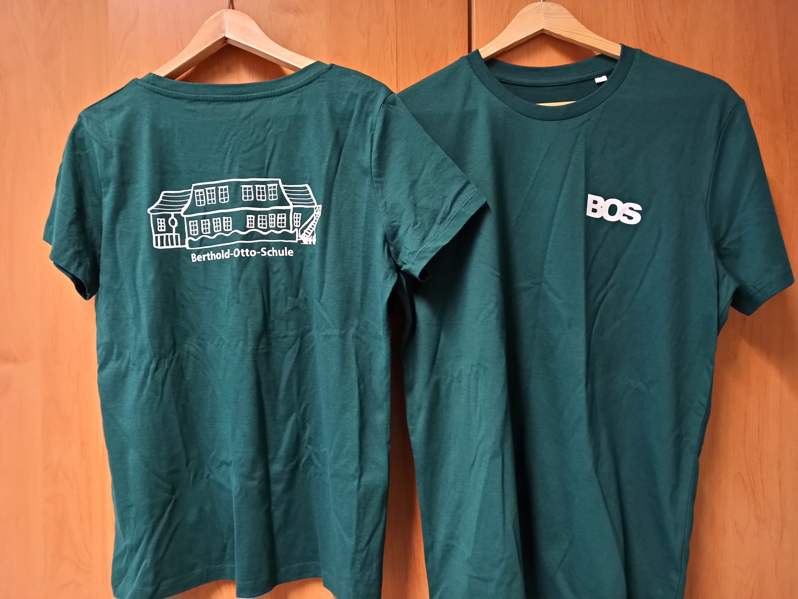 Ein dunkelgrünes T-Shirt mit den Buchstaben BOS auf der Vorderseite und dem gezeichneten Schulgebäude auf der Rückseite.